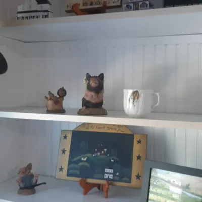Collection of rustic and country-style home decor displayed on white shelving, including pig figurines, folk art signs, miniature houses,...