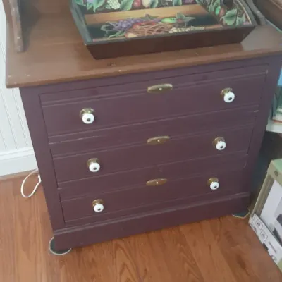Plum-painted wooden chest of drawers with white ceramic knobs and a natural wood gallery-top, featuring a decorative fruit-patterned tray...
