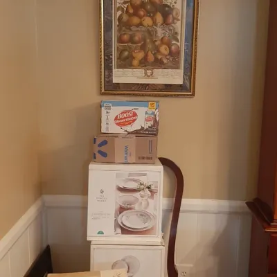 Framed botanical fruit print hanging above a stack of boxed household items and a striped upholstered dining chair.