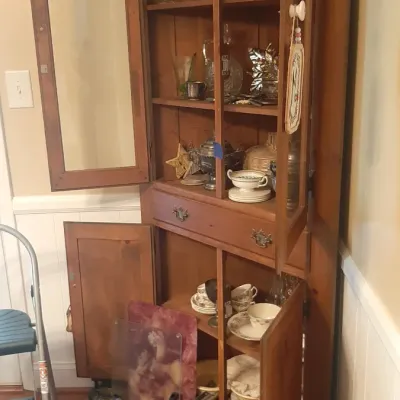 Vintage wooden corner hutch featuring glass-front upper cabinets, a center drawer, and lower storage cabinets filled with glassware,...