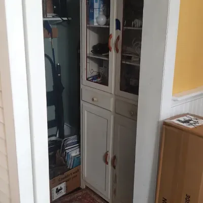 Tall white painted wooden cabinet with glass upper doors and reddish handles tucked into a storage room.