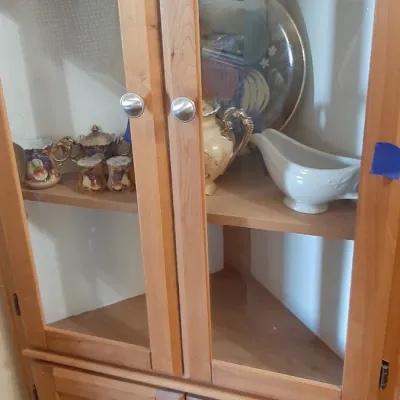 Light wood corner hutch with glass-front upper cabinet, lower wooden storage doors, and silver-tone knobs.