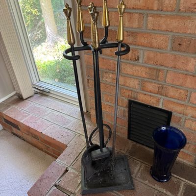 Vintage wrought iron fireplace tool set with brass finial handles, resting on a brick hearth beside a cobalt blue glass vase.