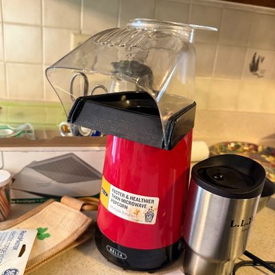 Bella red hot air popcorn popper with clear plastic chute, shown alongside a stainless steel L.L. Bean travel mug.