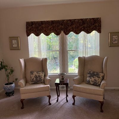 Pair of traditional beige upholstered wingback chairs with cabriole legs and pattern accent pillows, flanked by a small wooden side table.