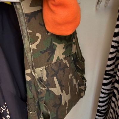 Camouflage hunting vest with front utility pockets hanging on a clothes rack next to a bright orange hat.