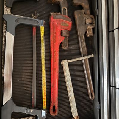 Assorted vintage hand tools arranged in a toolbox drawer, including a red Ridgid 18-inch heavy-duty pipe wrench, a hacksaw, and...
