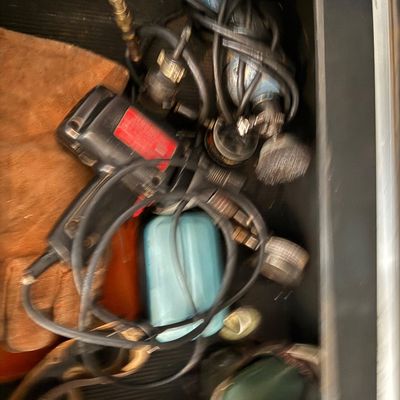 Assorted power tools and workshop accessories stored in a drawer, including a handheld drill, wire wheel attachment, protective gloves,...