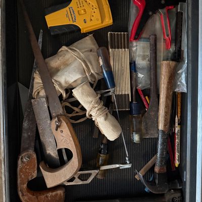 Assorted vintage hand tools in a toolbox drawer, including a Zircon stud finder, staple gun, claw hammer, saws, and folding ruler.