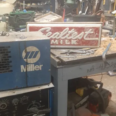 Workshop crowded with equipment including a Miller Syncrowave 250 arc welder, a vintage Sealtest Milk illuminated sign, tools, and...