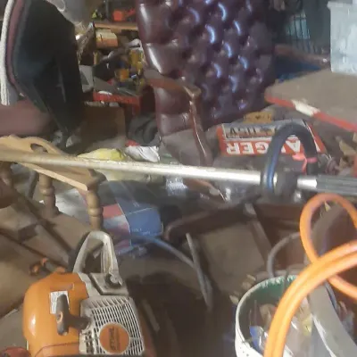 A cluttered workshop filled with various tools and equipment, including a Stihl chainsaw, string trimmer, and a burgundy tufted leather...