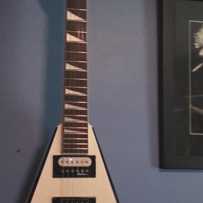 Sale Photo Thumbnail #14: Jackson Flying V electric guitar in white with black trim, featuring shark fin fretboard inlays and double humbucker pickups, displayed on a wall mount.