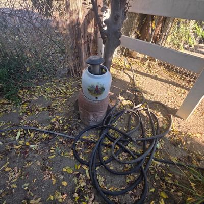 Sale Photo Thumbnail #72: Stack of outdoor garden items featuring an Olmec-style carved stone head pedestal, a ceramic rooster jar, and a solar garden light, set alongside a coiled garden hose.