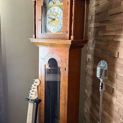 Sale Photo Thumbnail #83: Traditional wooden grandfather clock with moon phase dial, standing beside an EVH Frankenstein-style electric guitar on a stand and a vintage RCA microphone.