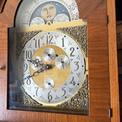 Sale Photo Thumbnail #84: Detail of a grandfather clock face featuring an ornate brass dial, moon phase display, and wooden case.