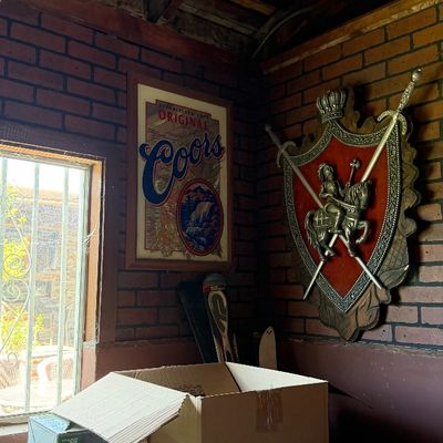 Sale Photo Thumbnail #87: Vintage framed Coors Original mirror sign paired with a large medieval-style decorative crest featuring crossed swords mounted on a faux-brick wall.