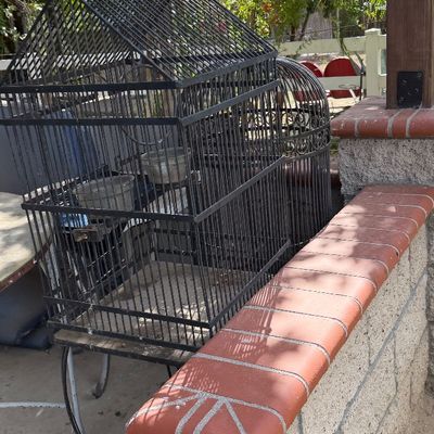 Sale Photo Thumbnail #90: Black metal house-shaped bird cage on a wrought iron stand, displayed outdoors alongside a second decorative cage.