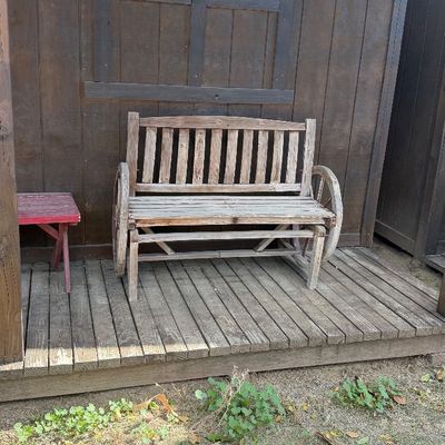 Sale Photo Thumbnail #95: Rustic weathered wooden garden bench with wagon wheel armrests, displayed alongside a small red wooden stool on a wooden porch.