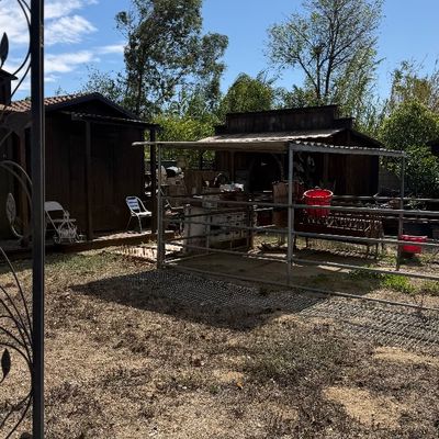 Sale Photo Thumbnail #98: Rustic outdoor yard setting with wooden storage sheds, metal fencing panels, and various farm and garden equipment under clear blue skies.