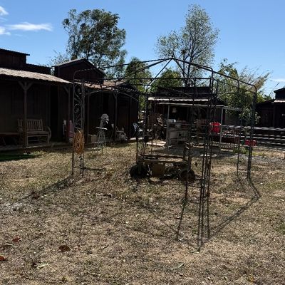 Sale Photo Thumbnail #100: Large metal gazebo structure standing in a grassy yard, featuring scrollwork details and a weathered, rusted finish.