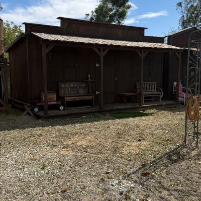 Sale Photo Thumbnail #101: Rustic Western-style wooden storage shed or playhouse featuring a covered front porch, false-front facade, and metal roof.