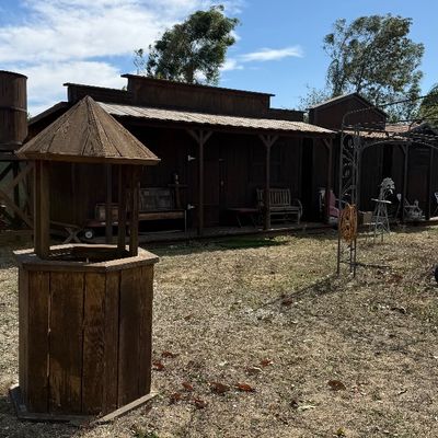 Sale Photo Thumbnail #105: Rustic wooden decorative wishing well structure situated in a yard featuring Western-style wooden buildings in the background.