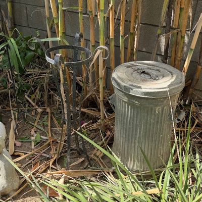 Sale Photo Thumbnail #117: Vintage metal trash can with lid alongside a black wrought iron plant stand, set outdoors against a block wall.