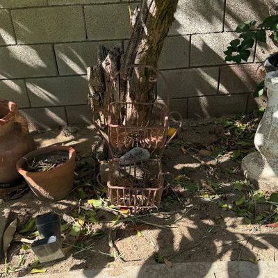 Sale Photo Thumbnail #122: Two-tier wrought iron wire basket planter stand positioned outdoors against a block wall, surrounded by terracotta pots.