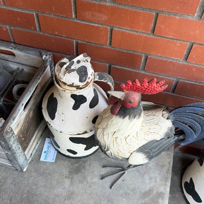 Sale Photo Thumbnail #130: Rustic metal rooster garden statue next to a painted cow-patterned milk can and a wooden crate against a red brick wall.