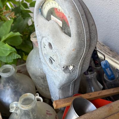 Sale Photo Thumbnail #132: Vintage cast metal parking meter head, showing an expired dial reading "No Pennies", surrounded by glass jugs in a wooden crate.