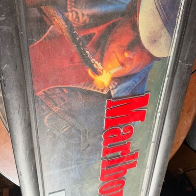 Sale Photo Thumbnail #152: Vintage framed Marlboro advertising sign featuring the Marlboro Man cowboy graphic, housed in a metallic frame.
