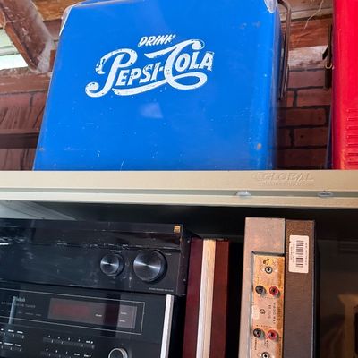 Sale Photo Thumbnail #154: Vintage blue metal Pepsi-Cola cooler resting on top of a metal shelf above various stereo receiver equipment.
