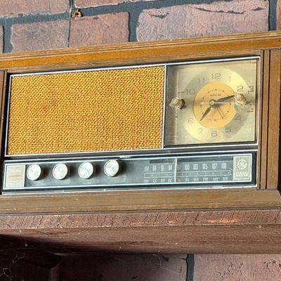 Sale Photo Thumbnail #165: Vintage General Electric wood-cased AM/FM clock radio with a fabric speaker grille and gold-tone dial face.