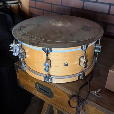 Sale Photo Thumbnail #167: Natural finish wooden snare drum with chrome hardware, featuring a Meinl cymbal resting on top and an accompanying drum key nearby.