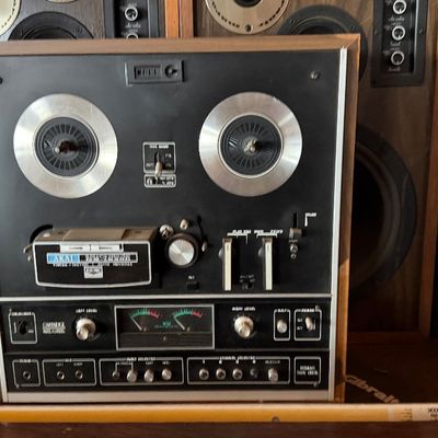 Sale Photo Thumbnail #168: Vintage Akai reel-to-reel tape deck featuring a black faceplate, silver knobs, and VU meters, housed in a wood cabinet alongside vintage audio speakers.