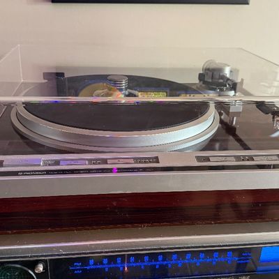 Sale Photo Thumbnail #196: Pioneer Quartz PLL direct drive automatic turntable with a dark wood grain base and clear dust cover, resting on an illuminated vintage stereo receiver.