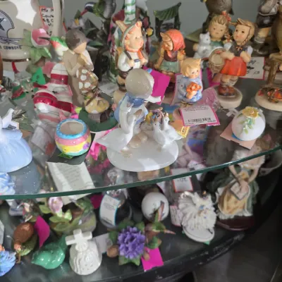 Sale Photo Thumbnail #61: Collection of vintage porcelain figurines, including Hummel-style children and bird statuettes, arranged on glass display shelves.