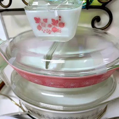 Sale Photo Thumbnail #169: Stack of vintage Pyrex glassware, including a small gooseberry pattern refrigerator dish and a pink Daisy pattern divided casserole dish with clear glass lids.