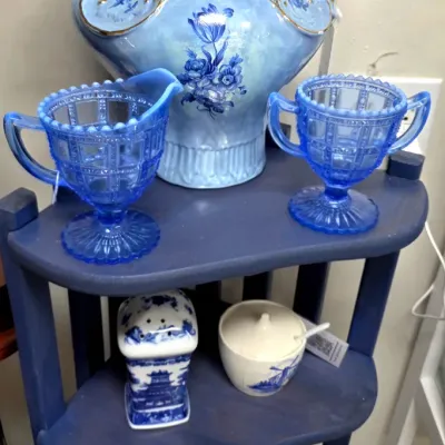Sale Photo Thumbnail #175: Collection of vintage blue glassware and porcelain display items, featuring a large iridescent blue water pitcher, matching press-cut creamer and sugar bowl, and blue-and-white ceramics on a blue corner shelf.