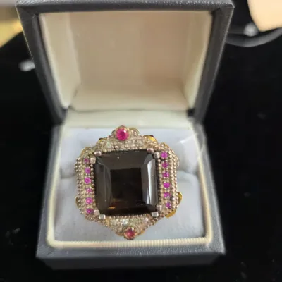 Sale Photo Thumbnail #290: Ornate statement ring featuring a large emerald-cut smoky quartz center stone surrounded by pink or red accent gems in an intricate silver and gold-tone setting, presented in a black jewelry box.