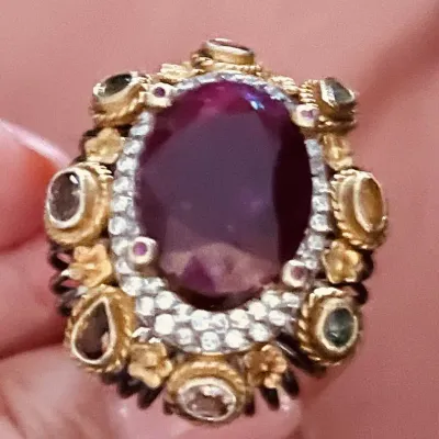 Sale Photo Thumbnail #355: Ornate statement ring featuring a central oval purple gemstone surrounded by pavé accents and textured gold-tone embellishments.