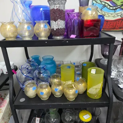 Sale Photo Thumbnail #420: Collection of decorative glass items arranged on a black three-tiered shelf unit, including frosted, gold-accented, blue, and purple vases and candle holders.