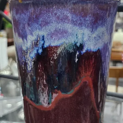 Sale Photo Thumbnail #491: Hand-thrown studio pottery tumbler or vase featuring a vivid multi-colored drip glaze in shades of purple, blue, and deep red with a natural tan rim.