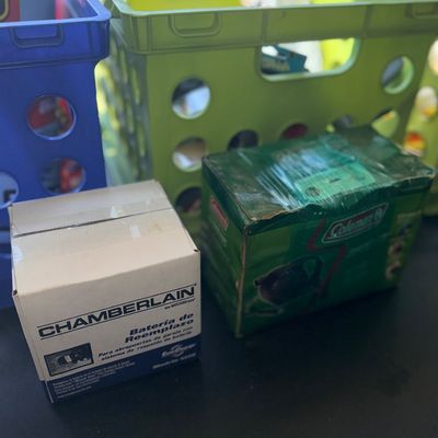 Sale Photo Thumbnail #94: Boxed Chamberlain replacement garage door battery and a Coleman item in original packaging, set in front of plastic storage crates.