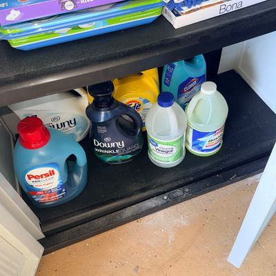 Sale Photo Thumbnail #120: Assorted household cleaning supplies and laundry products, including Persil, Downy, Tide, Clorox, and cleaning vinegar, arranged on black plastic shelving.