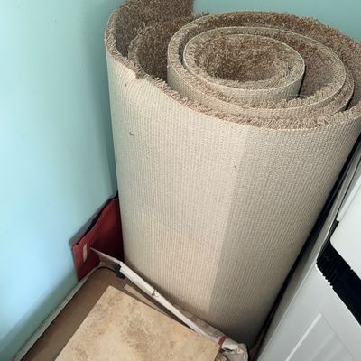 Sale Photo Thumbnail #128: Large roll of brown plush carpet upright in a corner alongside a box of flooring tiles.