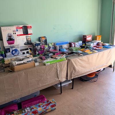 Sale Photo Thumbnail #173: Long folding tables covered with a variety of boxed household goods, electronics, lighting fixtures, and personal care items set up inside a garage.