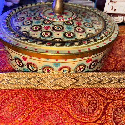 Sale Photo Thumbnail #340: Vintage gold-tone mesh watch band with a geometric diamond pattern, displayed alongside a decorative patterned metal tin on a red fabric surface.