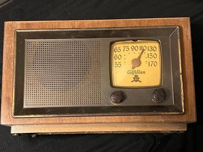 Vintage Gilfillan tabletop tube radio featuring a wooden cabinet, a metallic faceplate with a perforated speaker grille, and a yellowed...