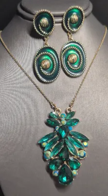 Sale Photo Thumbnail #116: A vintage costume jewelry set featuring a gold-tone chain necklace with a large, tiered pendant set with emerald green marquise and teardrop rhinestones, accented by iridescent aurora borealis rhinestones. Accompanied by a matching pair of gold-tone dangl